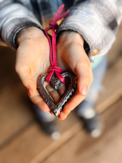 Heart-shaped blacksmith ornament with a red leather string held by child's hands against a blurred background